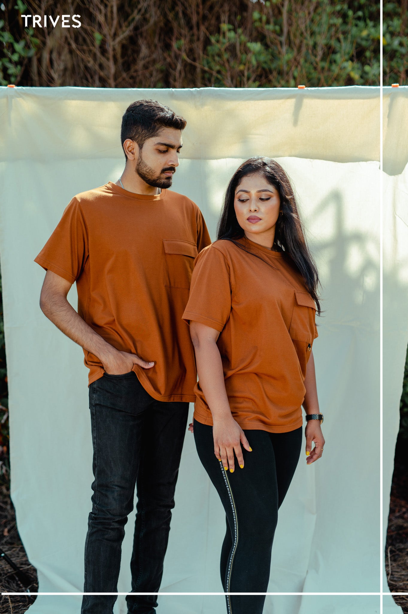 Two people wearing matching brown Trives cocoa Satash tee/t-shirts and black pants standing outdoors.