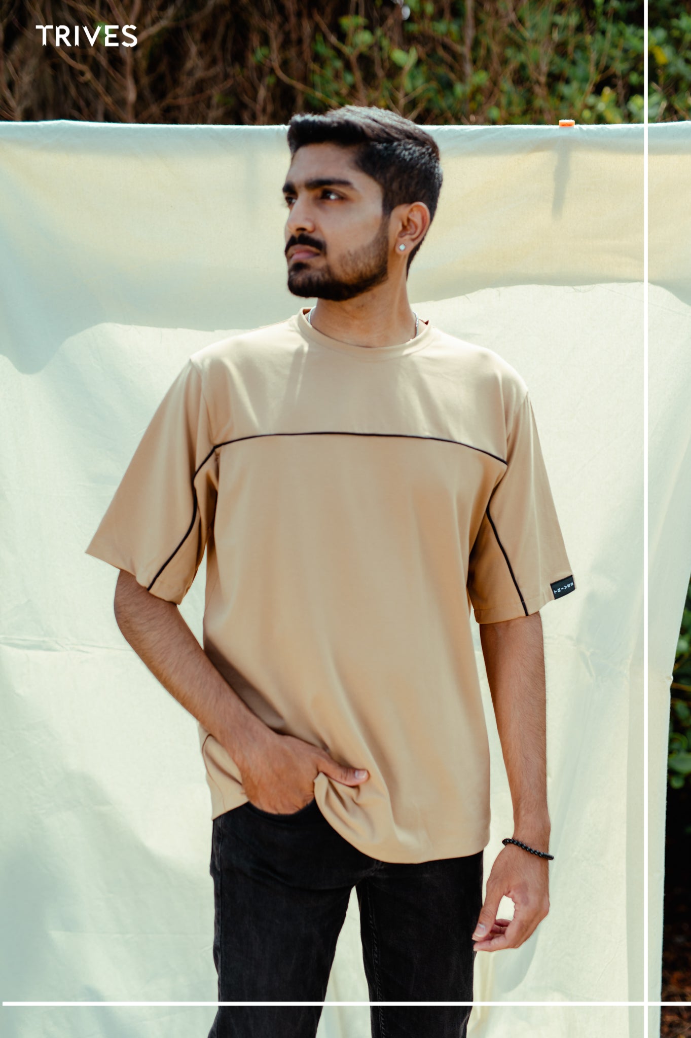 Man wearing a beige t-shirt with a TRIVES brand logo, standing outdoors.