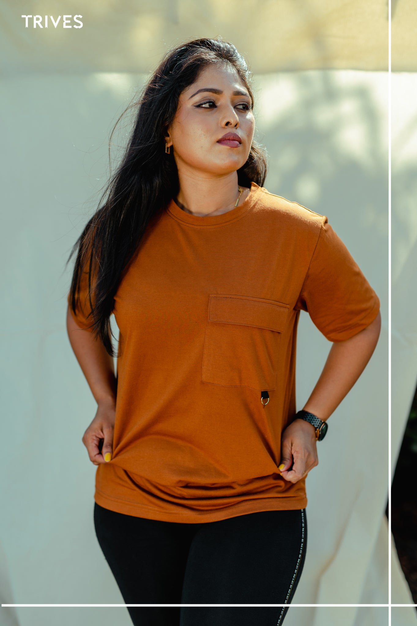 Woman wearing a trives cocoa/brown stash tee/ brown t-shirt with a pocket, standing outdoors.