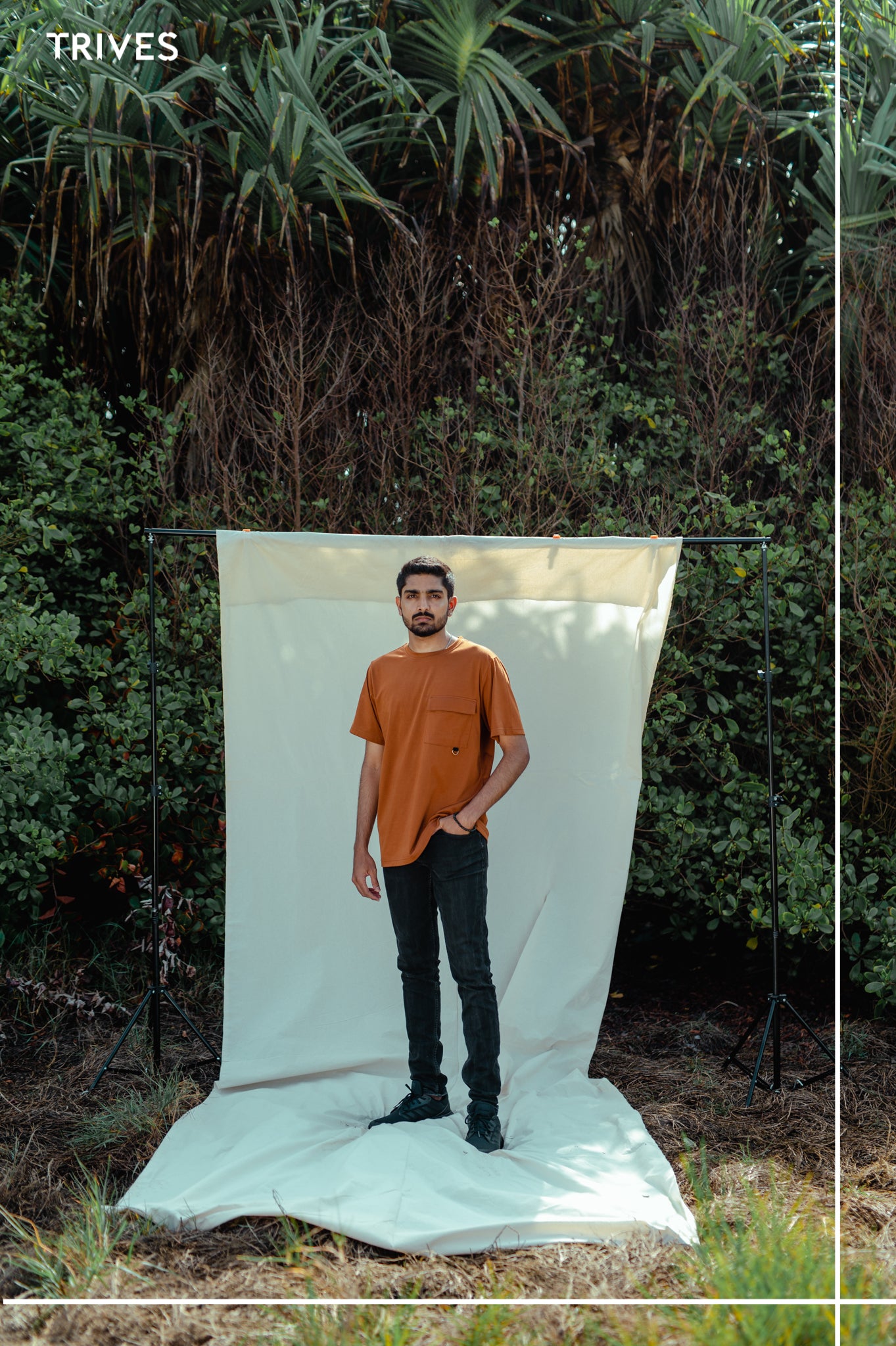 Person standing on a white sheet outdoors with greenery in the background. Trives Cocoa strash Tee