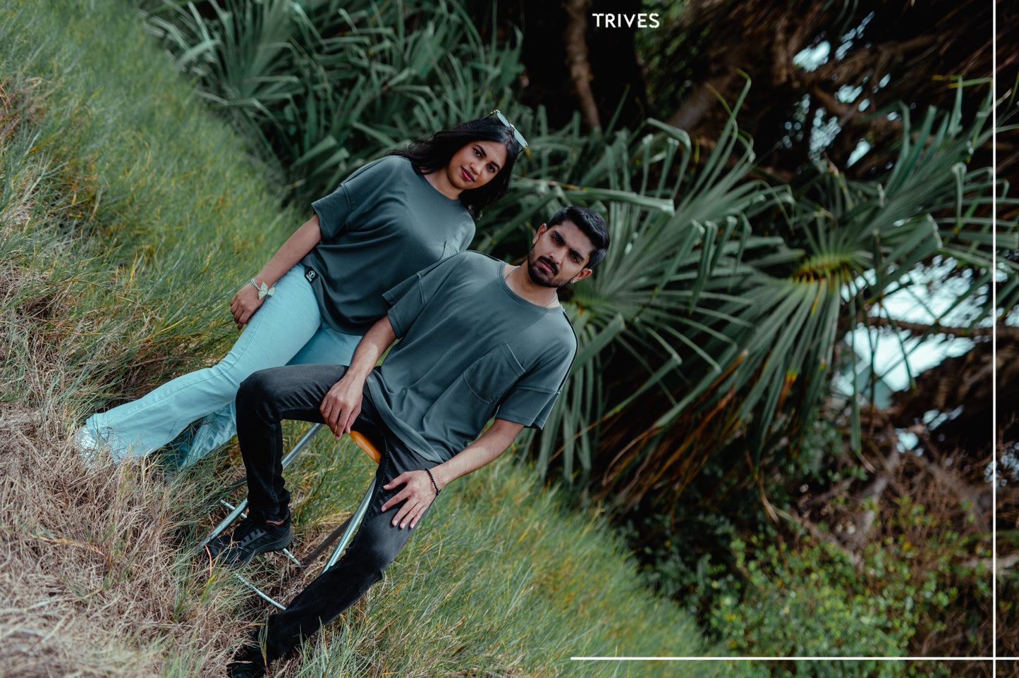 Two people sitting on a grassy hill with trees in the background, branded 'TRIVES'. Fray Tee is wearing