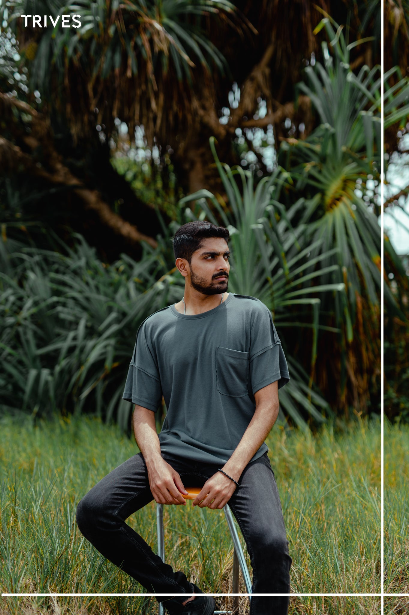 Man sitting on a stool in a grassy area with greenery in the background. Trives Fray Tee wearing