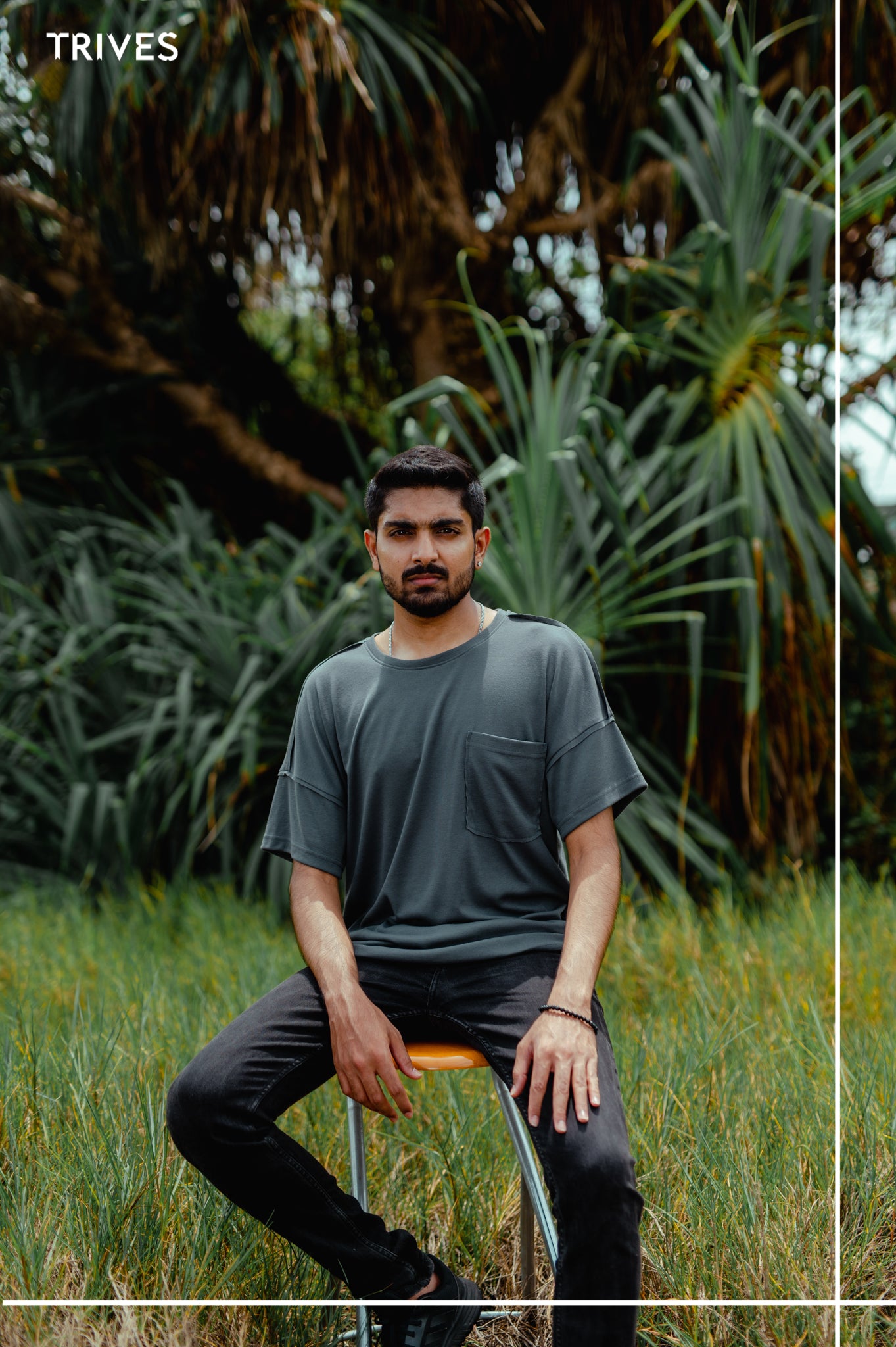 Man sitting on a stool in a grassy area with greenery in the background. Trives Fray Tee