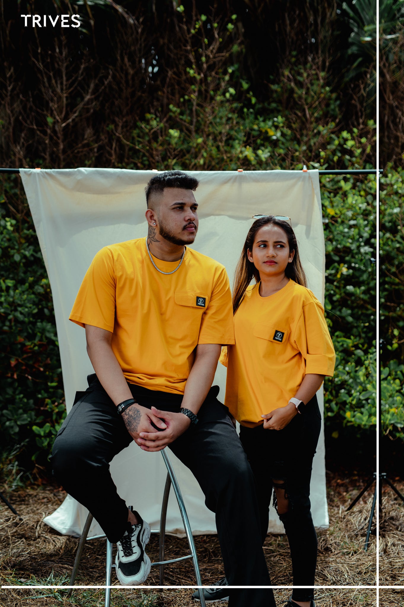 Two people wearing matching yellow t-shirts sitting outdoors with a white backdrop. Trives Vault Tee couple wearing
