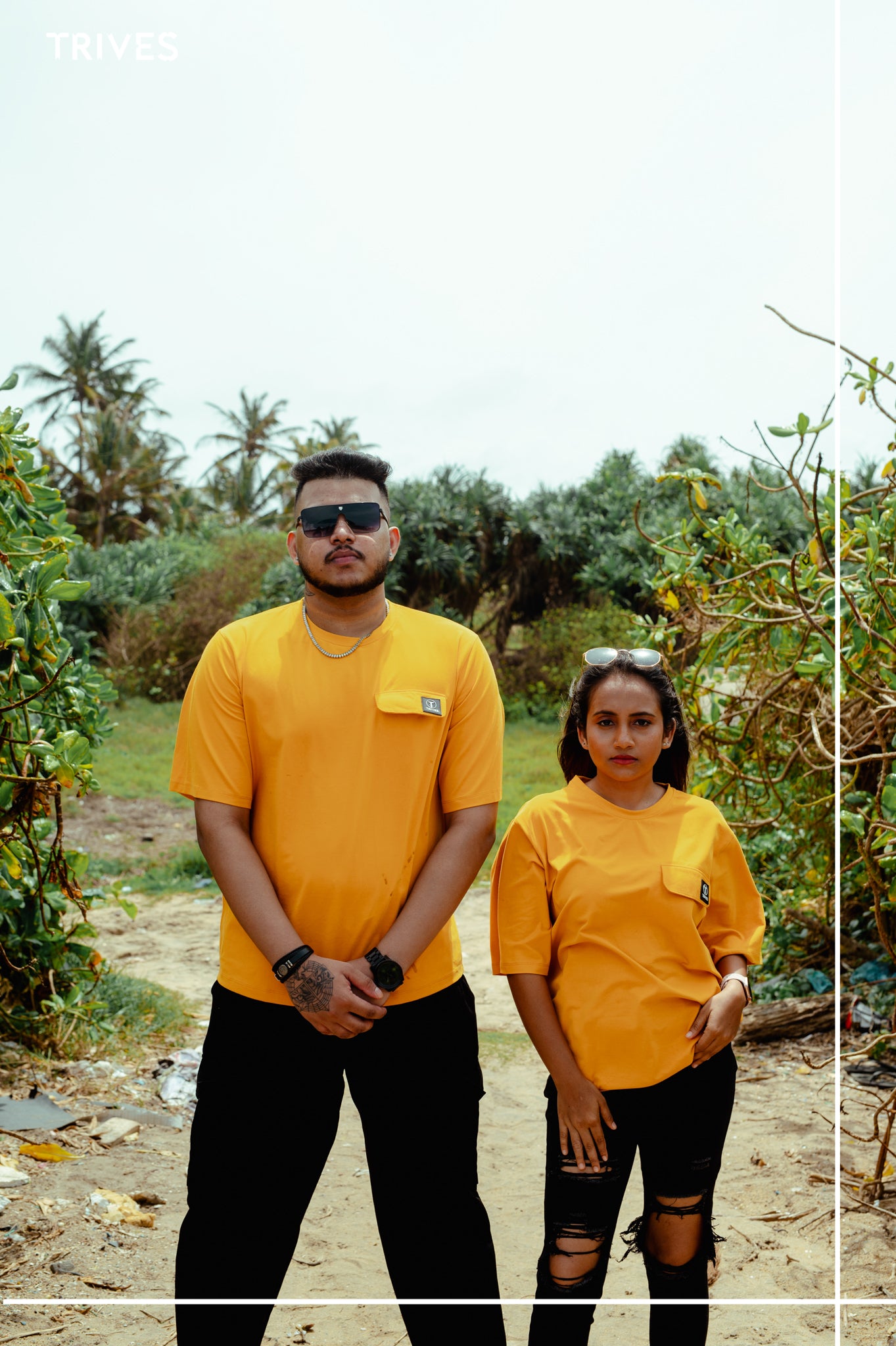 Two people wearing yellow shirts standing outdoors with greenery in the background. Trives Vault couple Tee