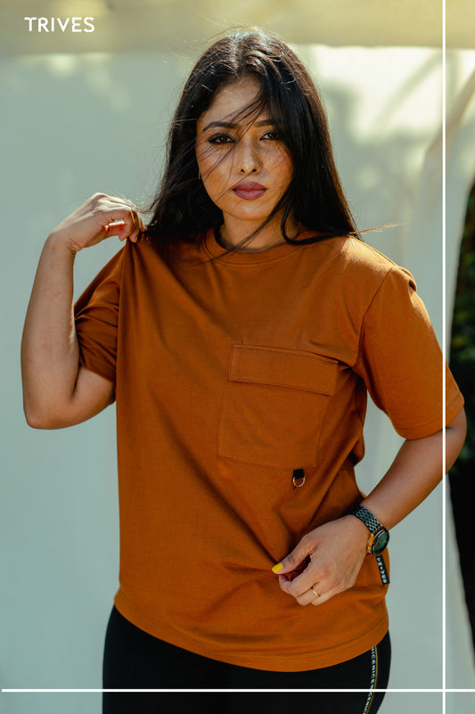 Woman wearing a brown TRIVES t-shirt with a visible brand logo, standing outdoors.