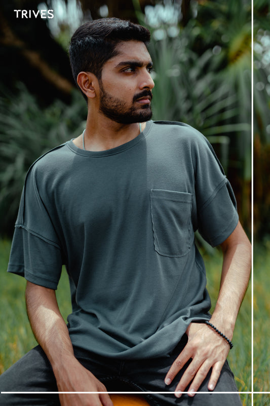Man wearing a light grey t-shirt sitting outdoors with greenery in the background. Trives Fray Tee 