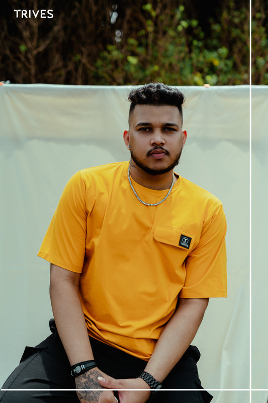 Man wearing a yellow t-shirt with a brand logo, sitting outdoors. Trives Vault Tee wearing. 