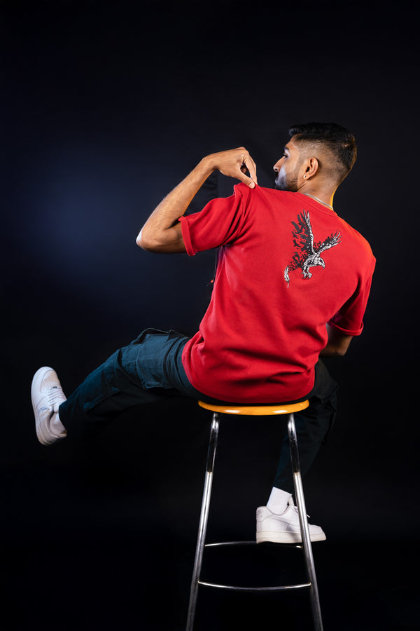 A person sitting on a stool wearing a red regular fit Trives t-shirt with an embroidered eagle design on the back.